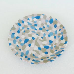 May include: A round, decorative plate with a speckled pattern in shades of blue, gray, and white. The plate has a light pink rim and is set against a white background. The design is reminiscent of terrazzo.