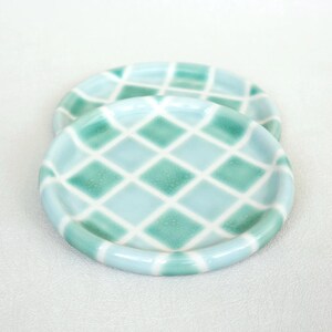 May include: Two small, round ceramic plates with a light blue and green diamond pattern. The plates have a raised rim and a glossy finish. The plates are on a white surface.