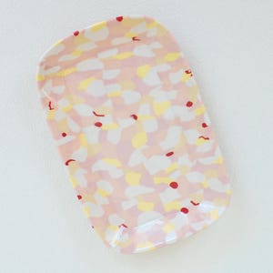 Könnte beinhalten: Ein rechteckiges Serviertablett aus Keramik mit abgerundeten Kanten. Das Tablett hat eine rosa Basis mit einem gesprenkelten Muster aus weißen, gelben und roten Formen. Das Tablett ist zum Servieren von Speisen oder zum Ausstellen von Dekorationsartikeln bestimmt.