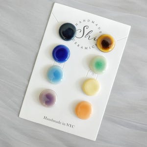 May include: A card of eight handmade ceramic buttons in various colours: black, blue, light blue, purple, brown, green, yellow, and orange. The card reads "Handmade in NYC" and "Shi Ceramics."