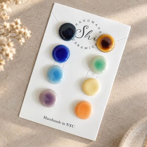 May include: A set of eight small, round, glossy ceramic buttons in various colors including dark blue, cobalt, light blue, lavender, amber, mint green, yellow, and orange, attached to a white display card labeled Handmade Shi Ceramics in NYC.