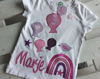 May include: White t-shirt with a pink, purple and white rainbow, a pink bird, and pink, purple and white balloons with a number 3 on one of them. The text "Marie" is embroidered in pink on the shirt.