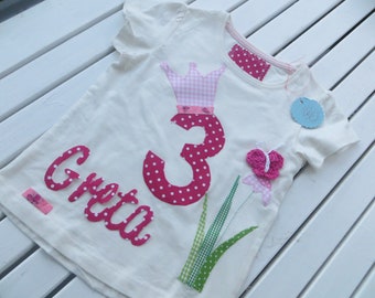 May include: White t-shirt with pink polka dot fabric appliques. The appliques spell out "Greta" and the number "3" with a pink crown and a pink butterfly.