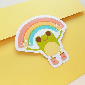 May include: A green frog with a big smile is holding up a rainbow with sparkling stars. The frog is wearing pink boots.