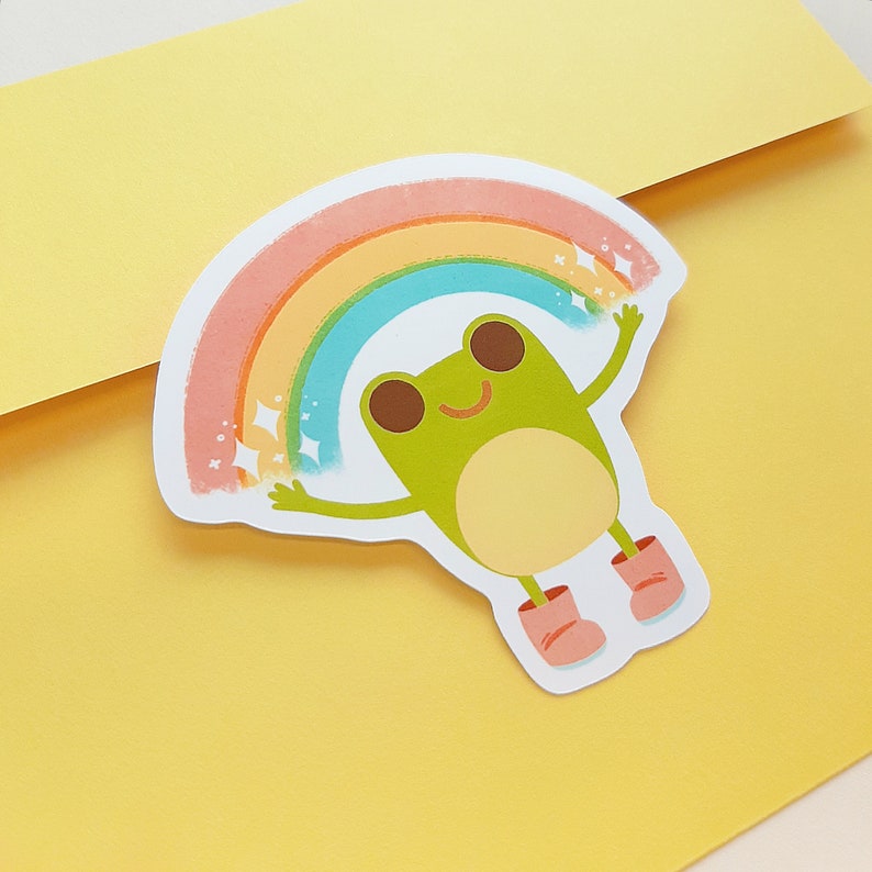 May include: A green frog with a big smile is holding up a rainbow with sparkling stars. The frog is wearing pink boots.