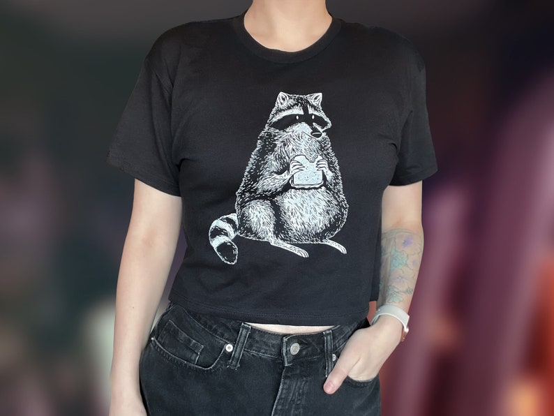 May include: A black cropped t-shirt with a white graphic of a raccoon eating a sandwich. The raccoon is wearing a mask and has a mischievous expression.