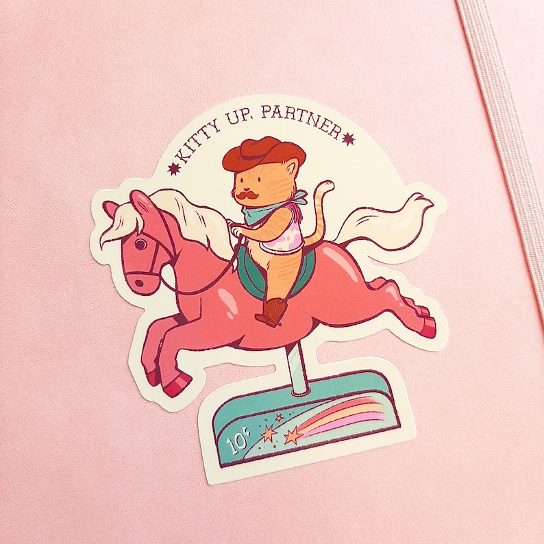 Op de afbeelding: Een roze draaimolenpaard met een kat die een cowboyhoed draagt en erop rijdt. Het draaimolenpaard heeft een witte manen en staart. De kat draagt een bandana en heeft een ster op zijn borst. De draaimolen heeft een bord met de tekst "Kitty Up, Partner" en "10&cent;".