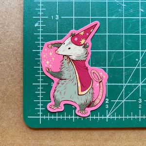May include: A pink sticker featuring a cartoon possum wearing a pink and gold wizard hat and a pink cape. The possum is casting a spell with its right paw, and there are sparkles around it.