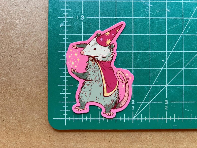 May include: A pink sticker featuring a cartoon possum wearing a pink and gold wizard hat and a pink cape. The possum is casting a spell with its right paw, and there are sparkles around it.