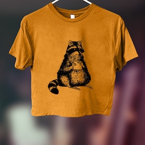 May include: A mustard yellow cropped t-shirt with a black and white illustration of a raccoon eating a sandwich.