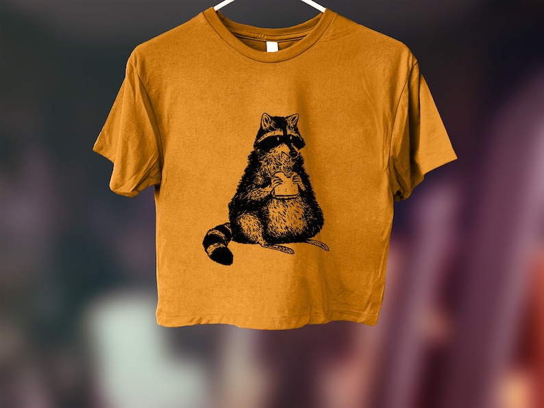 May include: A mustard yellow cropped t-shirt with a black and white illustration of a raccoon eating a sandwich.