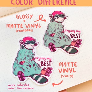 May include: Two raccoon stickers, one with a glossy finish and one with a matte finish. Both stickers feature a raccoon wearing a pink helmet and roller skates, with the text "crying my best". The glossy sticker has more saturated colors than the matte sticker.