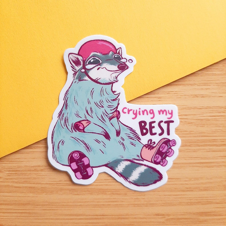 May include: A sticker featuring a cartoon raccoon wearing a pink hat and roller skates. The raccoon is sitting down with a sad expression and the text "crying my BEST" is written above it.