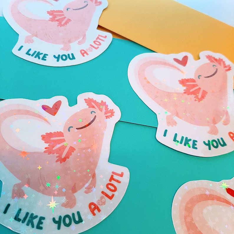 May include: Several holographic stickers featuring a smiling pink axolotl with a heart-shaped body and the text "I LIKE YOU A LOTL". The stickers have a rainbow design and are on a teal background.
