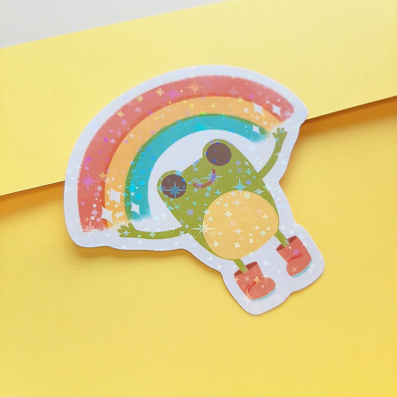 May include: A green frog wearing red boots is holding up a rainbow with glitter and stars. The frog is smiling and has big eyes.