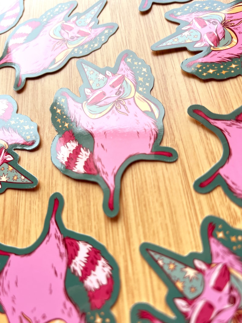 May include: A set of five pink raccoon stickers with a green background and stars. Each raccoon is wearing a wizard hat and has a pink and white striped tail.