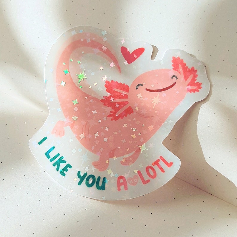 May include: A holographic sticker featuring a smiling pink axolotl in a heart shape. The sticker has the text "I like you a lotl" in teal and red. The axolotl has red gills and is decorated with star-shaped sparkles.