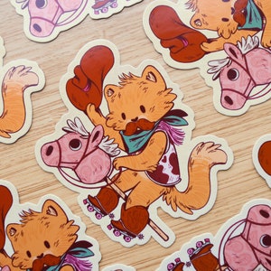 May include: A set of five colorful stickers featuring a cartoon cat wearing a cowboy hat and riding a pink rocking horse on roller skates. The cat has a brown mustache and is wearing a blue bandana and a brown and white cow print vest.