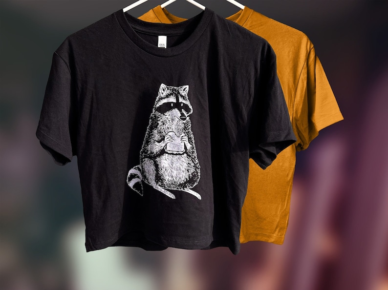May include: Black cropped t-shirt with a white graphic of a raccoon holding a sandwich.