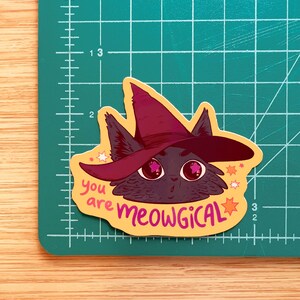 May include: A yellow sticker featuring a black cat wearing a purple witch hat. The cat has pink stars in its eyes and the sticker reads "you are meowgical" with pink stars surrounding the text.