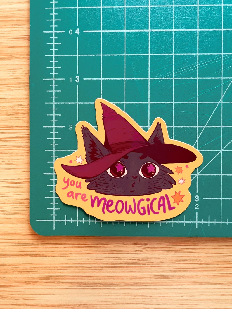 May include: A yellow sticker featuring a black cat wearing a purple witch hat. The cat has pink stars in its eyes and the sticker reads "you are meowgical" with pink stars surrounding the text.