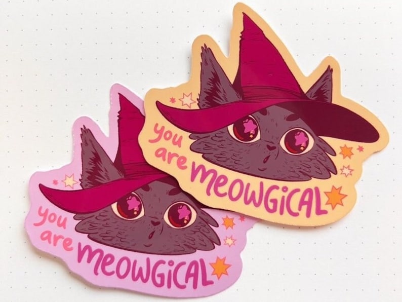 May include: Two pink and orange stickers featuring a cartoon cat wearing a pink witch hat. The cat has large, pink eyes and a surprised expression. The text on the stickers reads "you are meowgical".