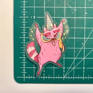 May include: A pink raccoon wearing a blue and white pointed hat with a yellow cape and stars around it. The raccoon is standing on its hind legs with its arms raised in the air.