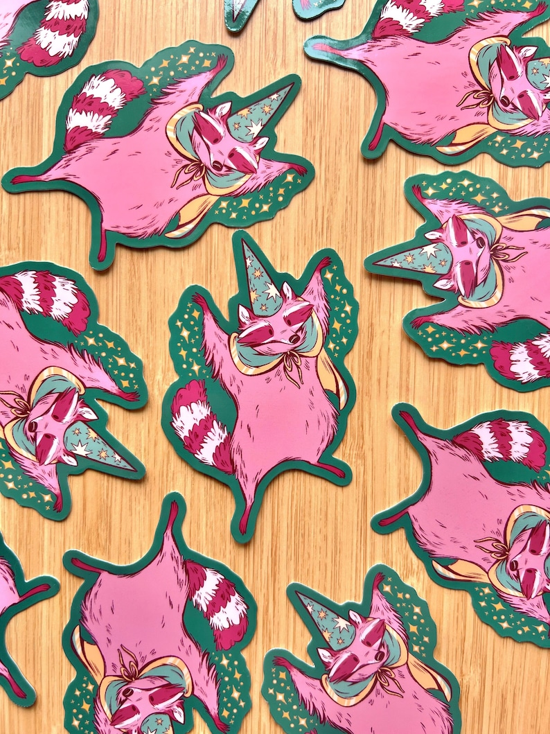 May include: A set of nine pink raccoon stickers with a green background and gold stars. Each raccoon is wearing a blue and green wizard hat and a yellow cape. The raccoons are in various poses, some with their arms raised and some with their arms down.