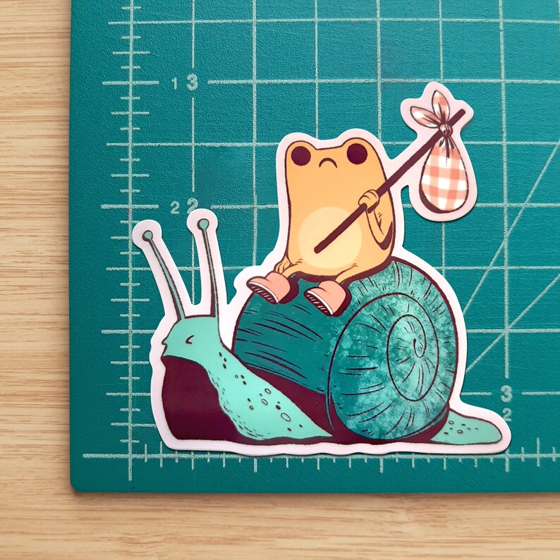 May include: A green snail with a yellow frog riding on its back. The frog is holding a stick with a red and white checkered bag attached to it. The snail is on a white background.
