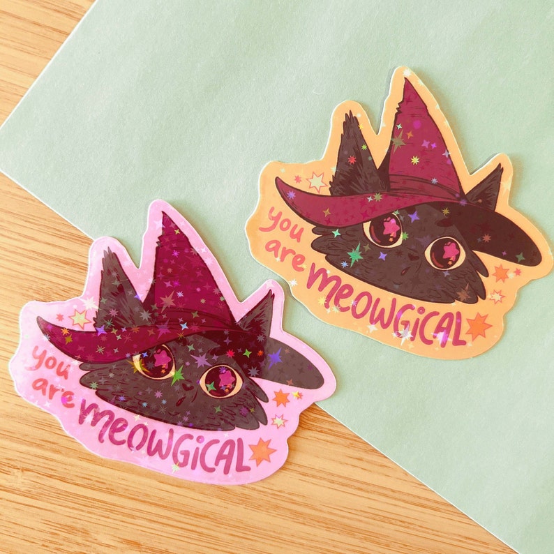 May include: Two pink and orange stickers featuring a black cat wearing a purple and pink witch hat. The cat has large, round eyes and is surrounded by glitter and stars. The text on the stickers reads "You are meowgical".