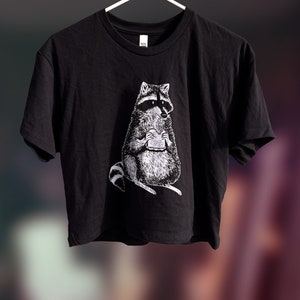 May include: A black cropped t-shirt with a white graphic of a raccoon holding a sandwich.