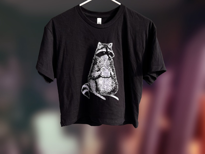 May include: A black cropped t-shirt with a white graphic of a raccoon holding a sandwich.