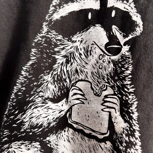 May include: A black and white illustration of a raccoon eating a sandwich. The raccoon is drawn in a cartoon style with a mischievous expression.