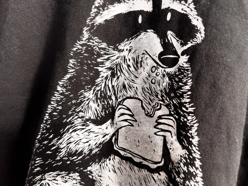 May include: A black and white illustration of a raccoon eating a sandwich. The raccoon is drawn in a cartoon style with a mischievous expression.