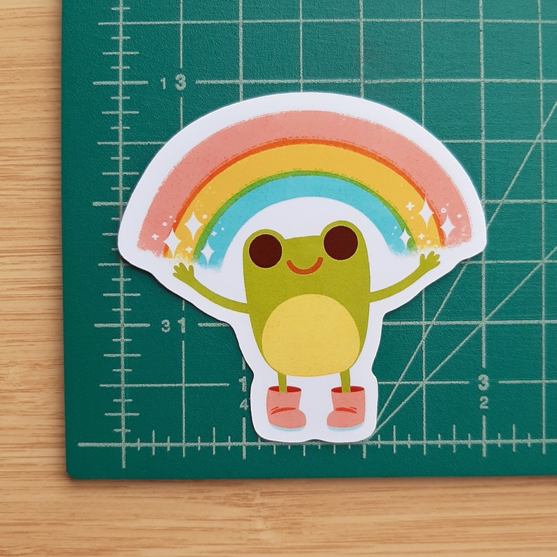 May include: A green frog with a big smile is holding up a rainbow with pink, yellow, and blue stripes. The rainbow has sparkling stars on each side.