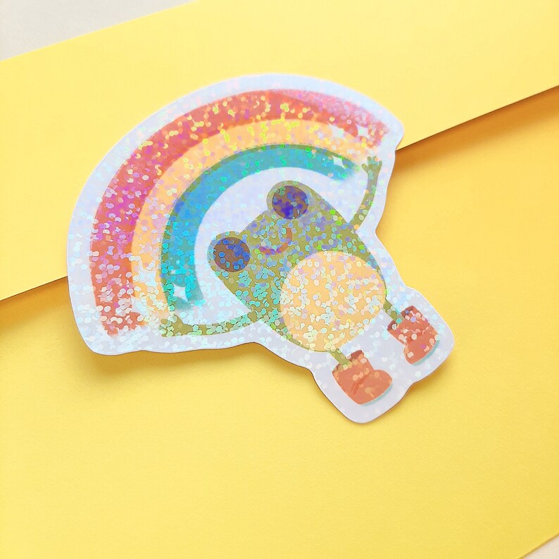 May include: A holographic sticker of a green frog holding up a rainbow with glitter accents.