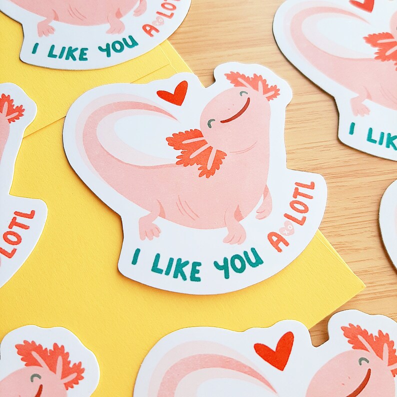 May include: A collection of stickers featuring a smiling pink axolotl with orange gills and the text "I LIKE YOU A LOTL". Each sticker has a heart and a rainbow design. The stickers are on a yellow envelope and a wooden surface.