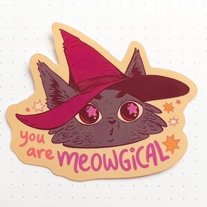 May include: A sticker featuring a cartoon cat wearing a pink witch hat with stars around it. The text "you are meowgical" is written below the cat.