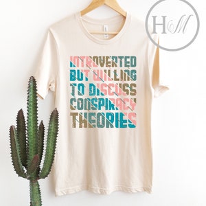 May include: A beige t-shirt with the text "Introverted but willing to discuss conspiracy theories" printed in a retro style with colorful stripes.