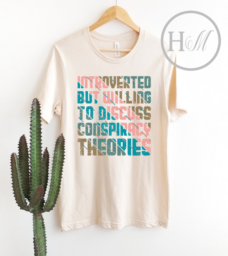 May include: A beige t-shirt with the text "Introverted but willing to discuss conspiracy theories" printed in a retro style with colorful stripes.