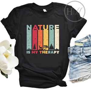 Puede incluir: Camiseta negra con un gr&aacute;fico de estilo vintage que dice "NATURE IS MY THERAPY". El gr&aacute;fico presenta un arco&iacute;ris de colores y una silueta de &aacute;rboles, un ciervo y una fogata.