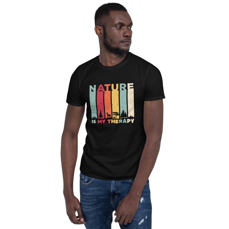 Puede incluir: Una camiseta negra con un gr&aacute;fico de estilo vintage que dice "NATURE IS MY THERAPY". El gr&aacute;fico presenta un fondo a rayas de colores con siluetas de &aacute;rboles y un ciervo.
