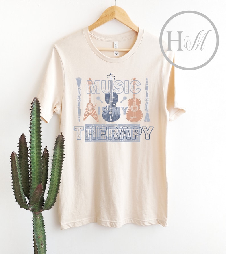 Pode incluir: Uma camiseta bege com o texto "Music is my therapy" estampado na frente. O texto &eacute; cercado por imagens de instrumentos musicais, incluindo uma guitarra, um violino, um clarinete e bateria.