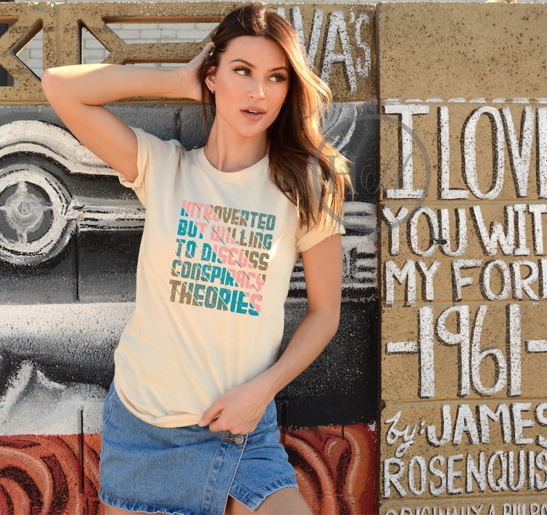 May include: A woman wearing a light beige t-shirt with the text "Introverted but willing to discuss conspiracy theories" printed in pink, blue, and teal. The text is stacked vertically and the shirt is a relaxed fit.
