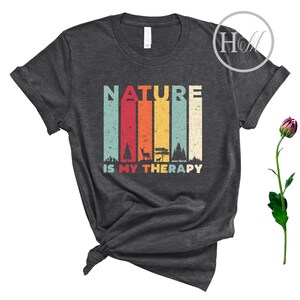 Puede incluir: Una camiseta gris oscuro con un gr&aacute;fico de estilo vintage que dice "NATURE IS MY THERAPY". El gr&aacute;fico presenta un fondo a rayas de colores con im&aacute;genes de &aacute;rboles, un ciervo y una tienda de campa&ntilde;a.