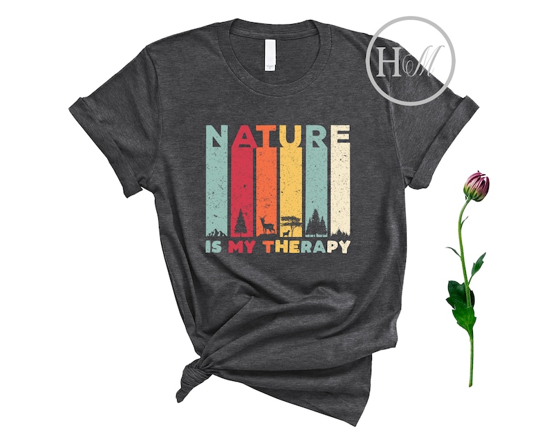 Puede incluir: Una camiseta gris oscuro con un gr&aacute;fico de estilo vintage que dice "NATURE IS MY THERAPY". El gr&aacute;fico presenta un fondo a rayas de colores con im&aacute;genes de &aacute;rboles, un ciervo y una tienda de campa&ntilde;a.