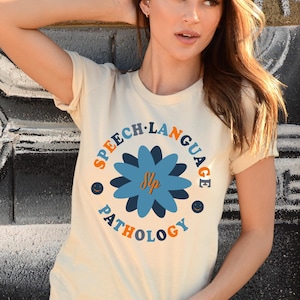 May include: A beige t-shirt with a blue flower graphic and the text "Speech-Language Pathology" in a colorful circle around the flower. The text "SLP" is inside the flower.