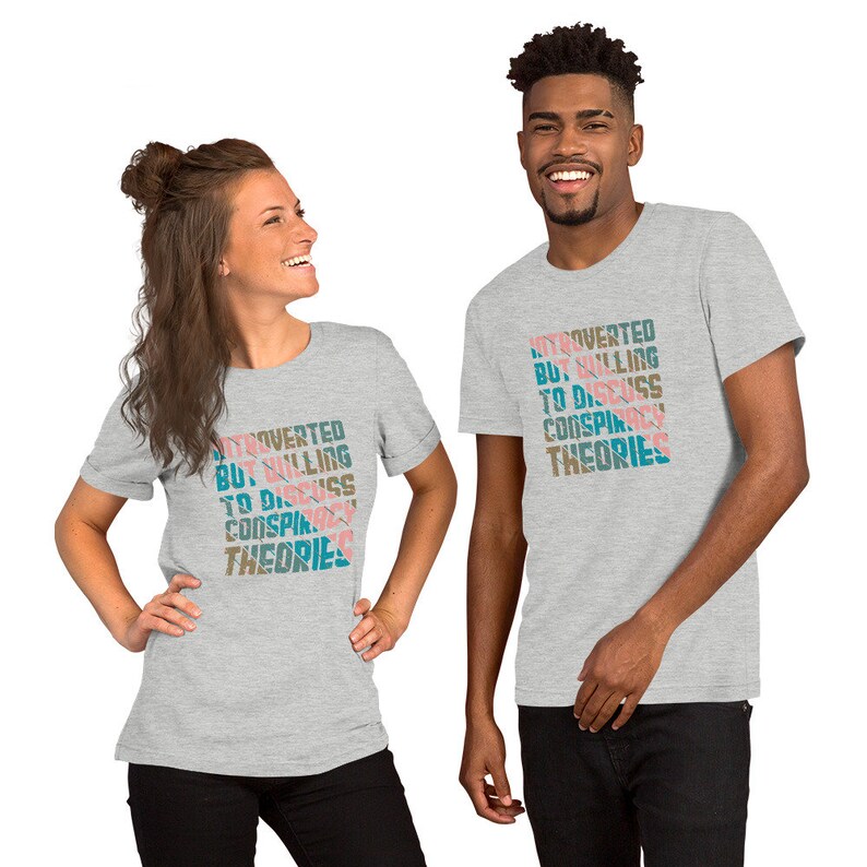 May include: Two people wearing gray t-shirts with the text "Introverted but willing to discuss conspiracy theories" printed in a colorful, layered font.