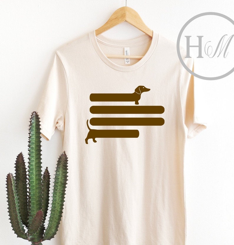 May include: A beige t-shirt with a brown dachshund silhouette design. The dachshund is depicted walking across four horizontal brown lines.