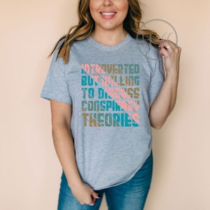 May include: A gray t-shirt with a graphic design that reads "Introverted but willing to discuss conspiracy theories". The text is in a colorful, striped font.